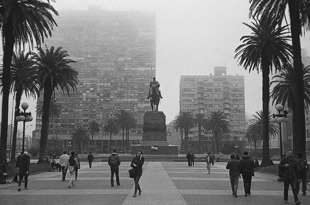Praça com árvores de palmeira, pessoas caminhando, estátua de um cavaleiro ao centro e prédios altos ao fundo em um dia nublado.