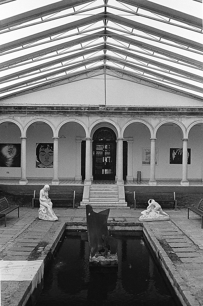Interior de uma galeria de arte com teto de vidro, pilares arqueados e duas esculturas de pessoas sentadas ao lado de uma fonte central.