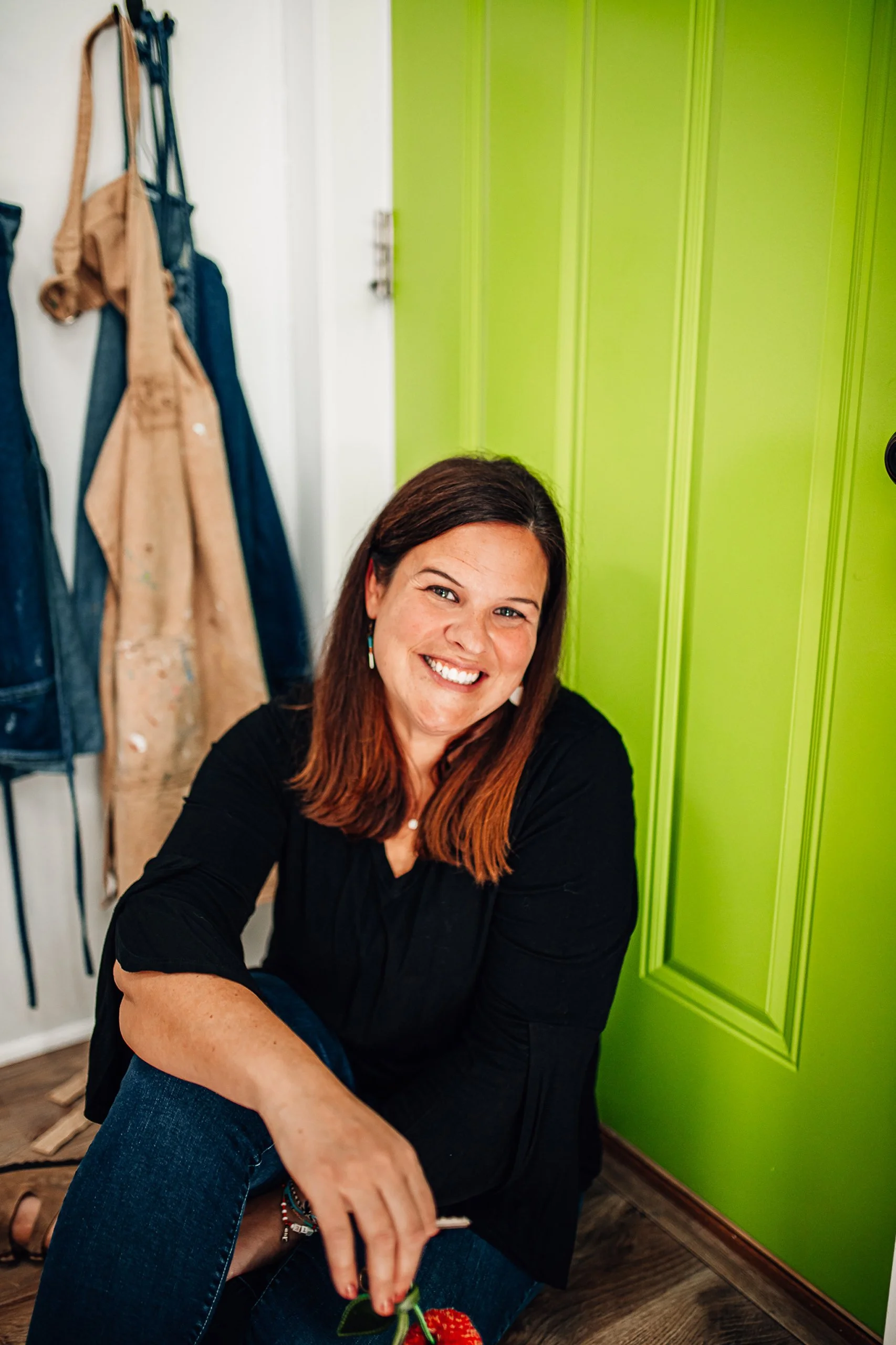 A woman with brown hair and a black top smiling while sitting on the floor next to a bright green door.