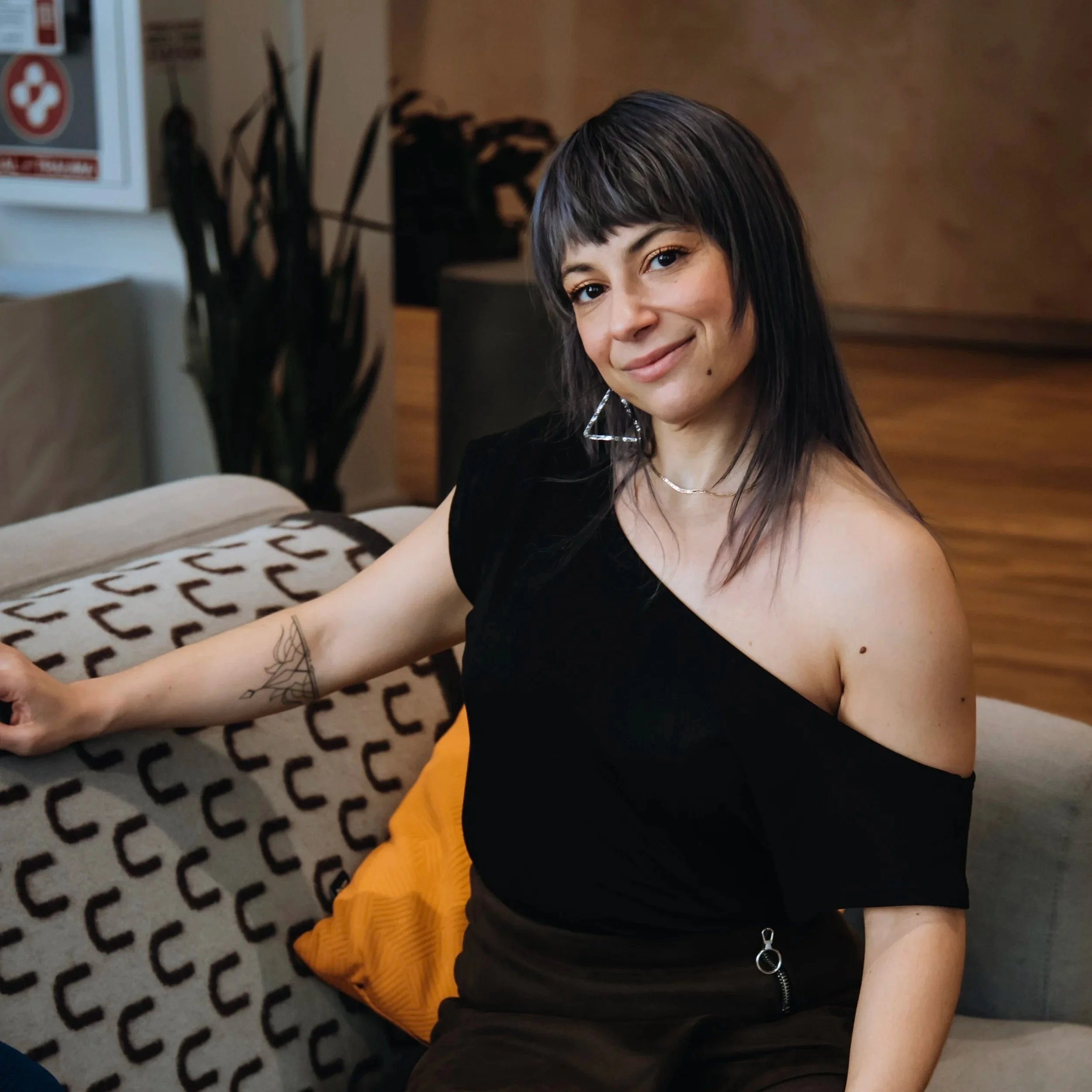 A young woman with dark hair and a tattoo on her left arm, wearing a black off-the-shoulder top and yellow skirt, sitting on a beige sofa in a room with wooden paneling, smiling at the camera.
