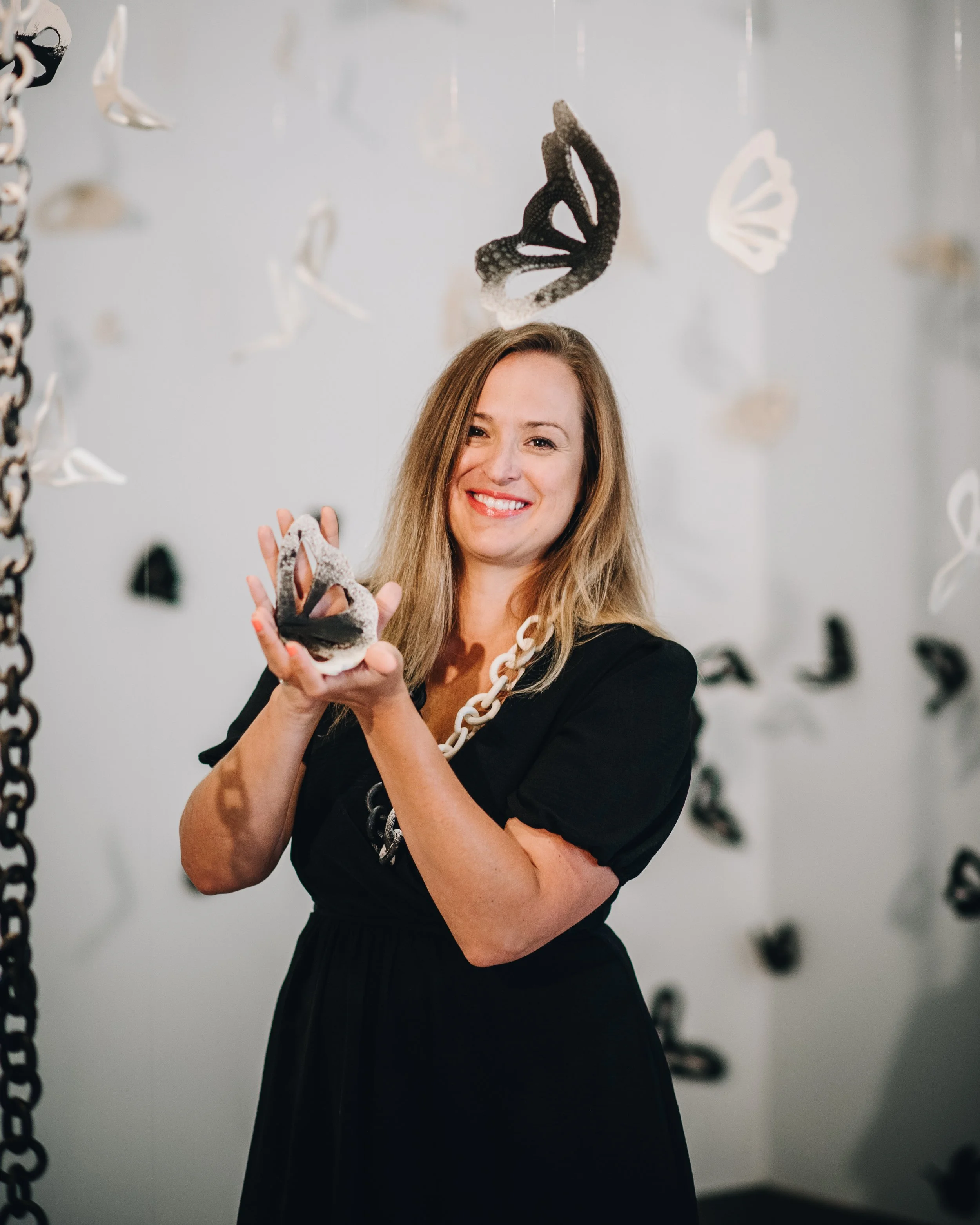 A woman with long blonde hair wearing a black dress and a white chain necklace, smiling while holding a butterfly-shaped sculpture in a gallery with butterfly art hanging from the ceiling.