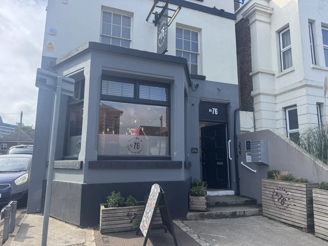 Exterior of a coffee shop at 76, with large front window, black door, and wooden planters outside, located in a multi-story building.