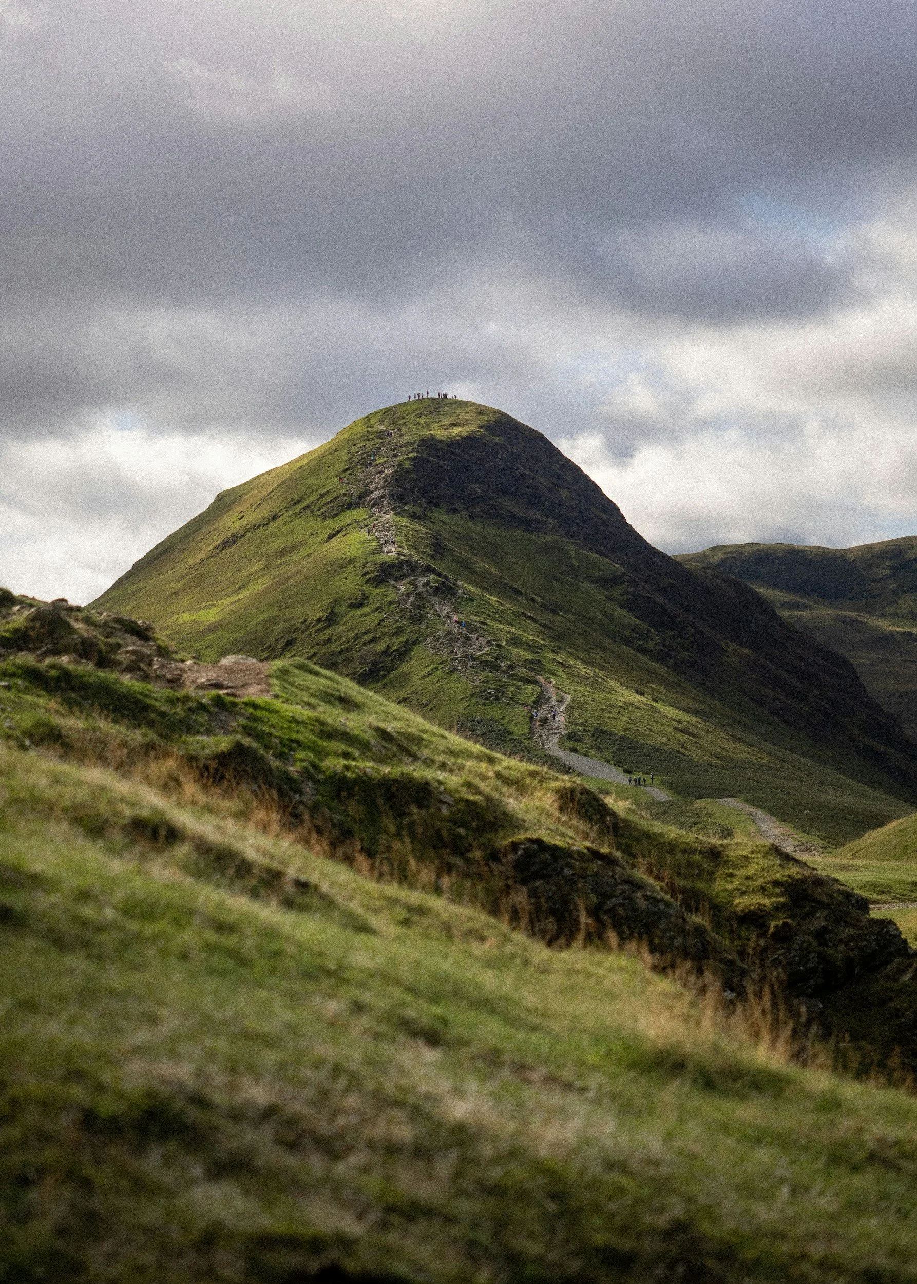 A tall green mountain with a trail leading up to the top, under a cloudy sky. A line of hikers can be seen near the summit.