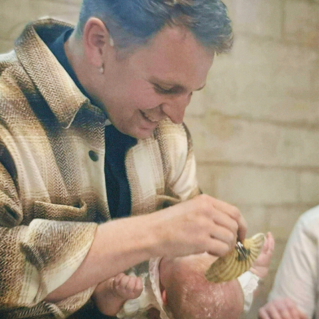 A woman smiling while holding a shell ornament near a large round pumpkin, indoors with a wooden wall background.