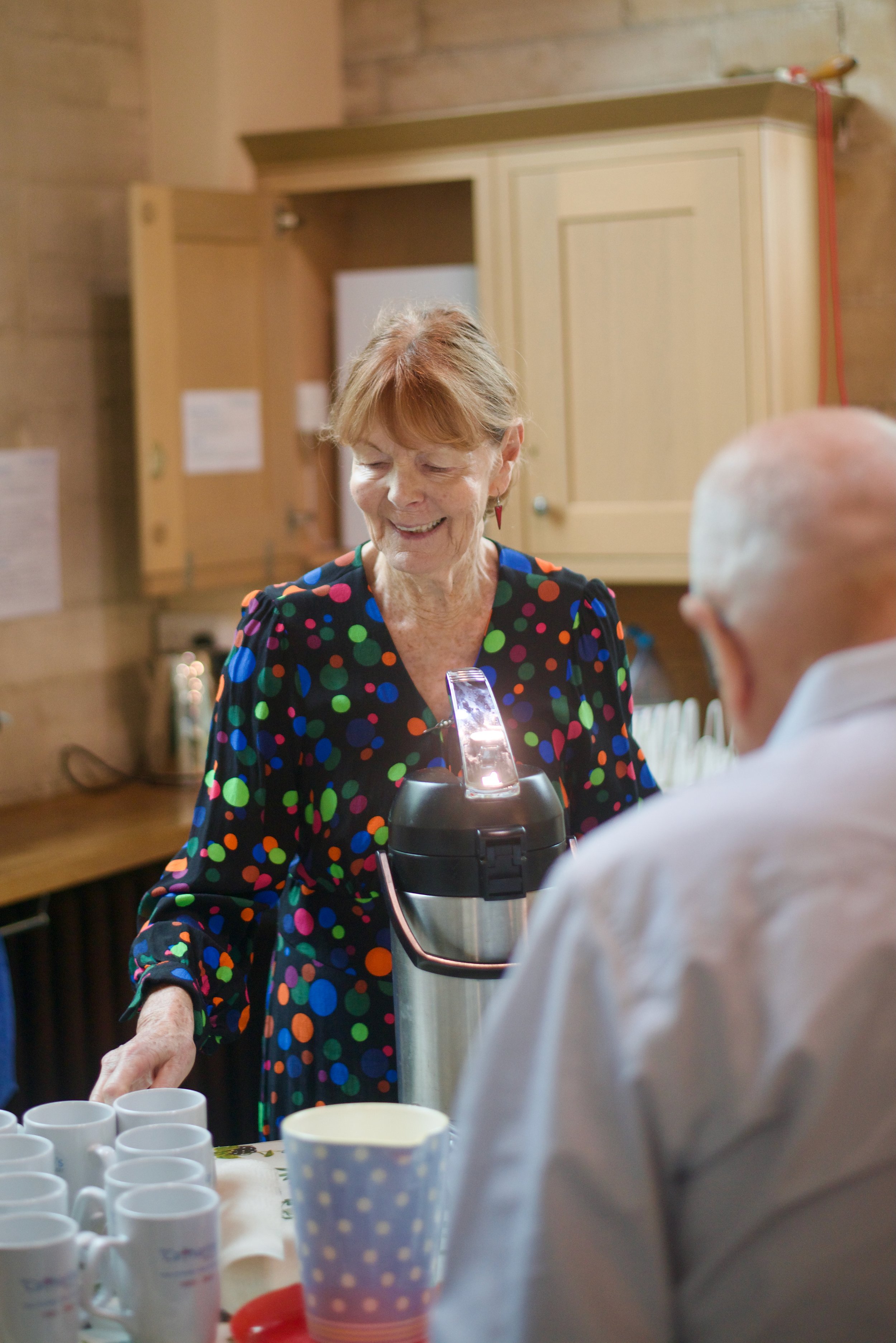 A smiling elderly woman wearing a black dress with colorful polka dots serves hot drinks to an elderly man at a gathering, with multiple coffee mugs on the table.