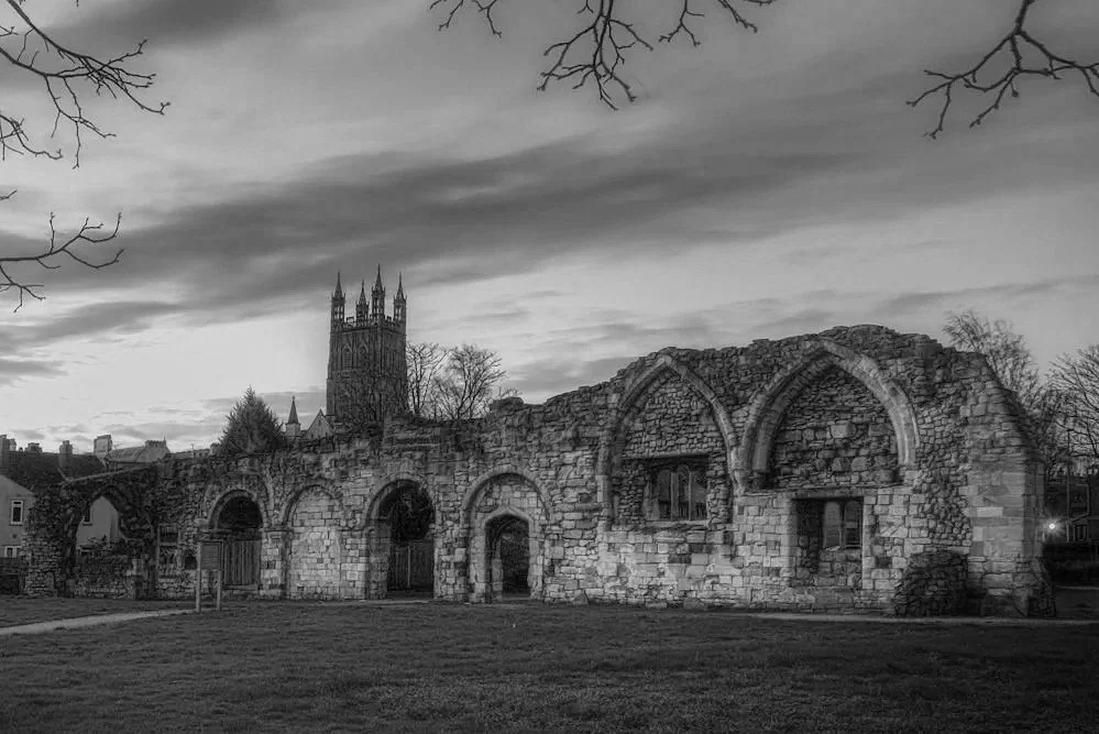 Black and white photograph of an old stone church ruins with Gothic architecture, featuring pointed arches, small windows, and a tall church tower in the background, with leafless trees and a cloudy sky.