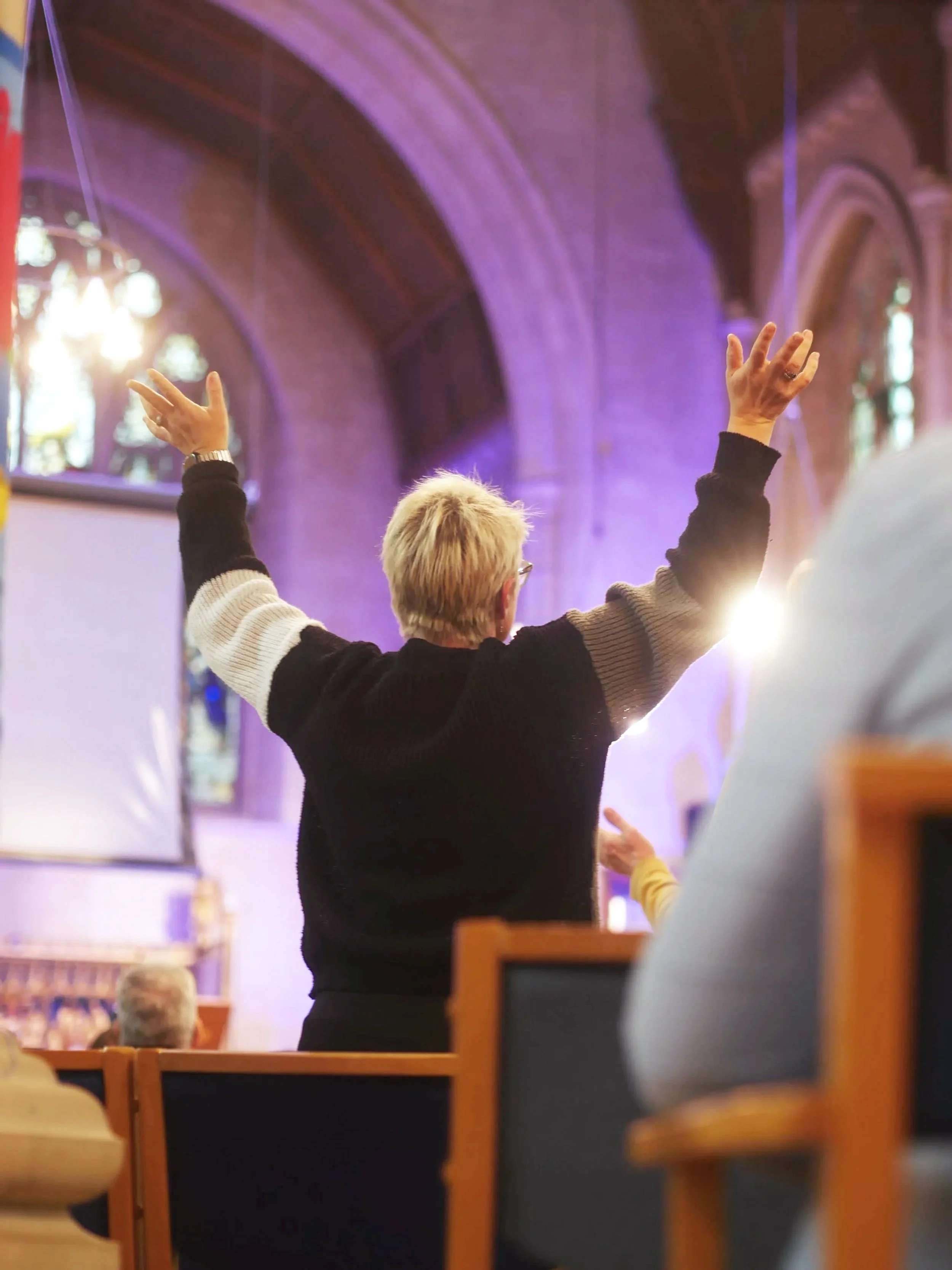 A person with short blonde hair and glasses standing in a church or cathedral with arms raised, facing a large screen, during a church service or event.