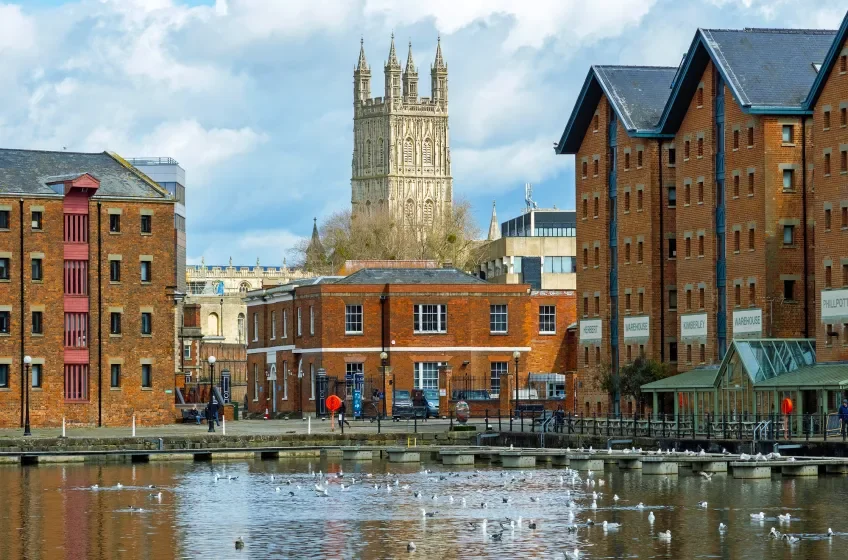 A cityscape featuring brick buildings, a body of water with birds, and a tall historic stone tower in the background.