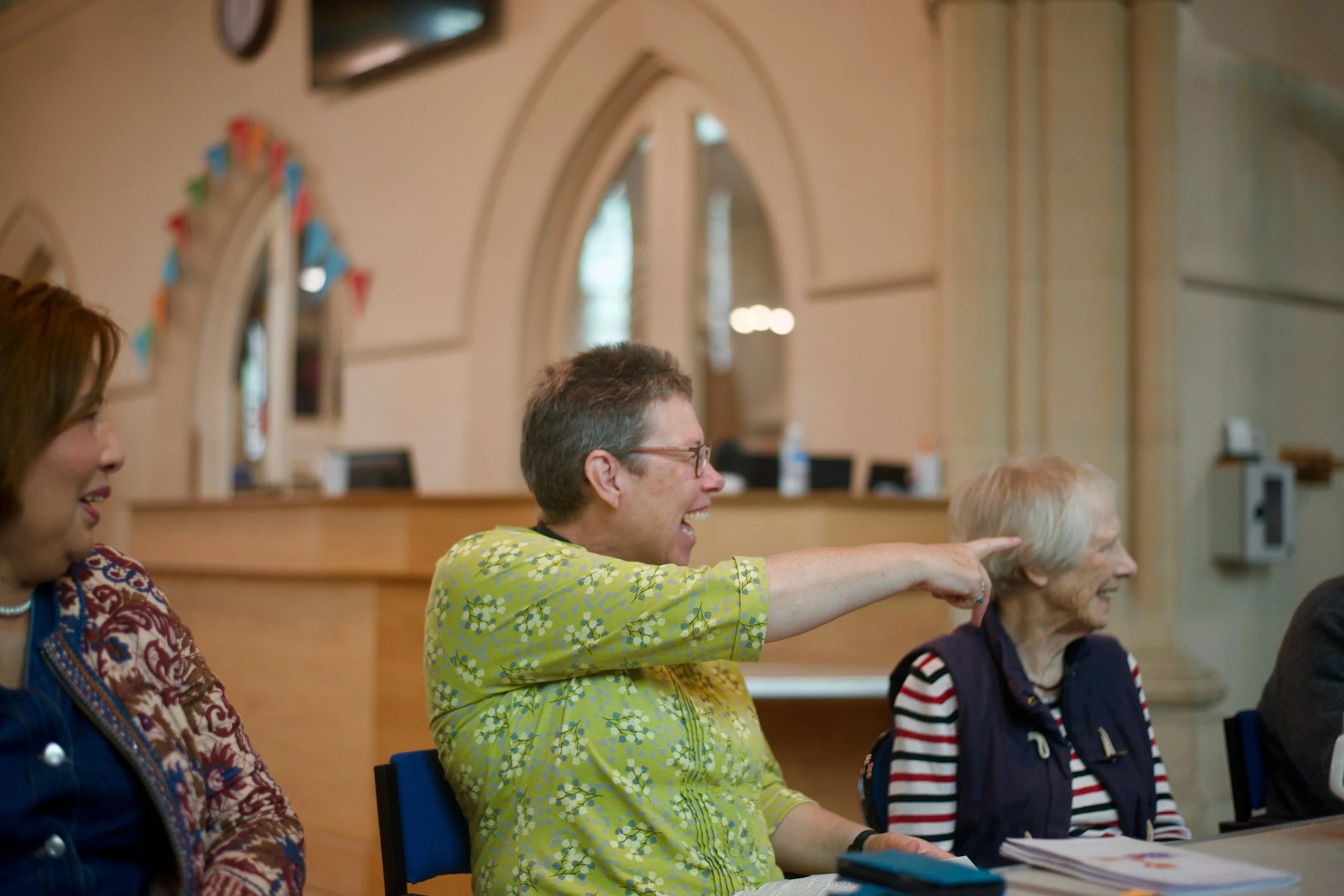 A group of diverse women, with one woman playfully touching another's head, sitting at a table in a room with arched windows and wooden decor.