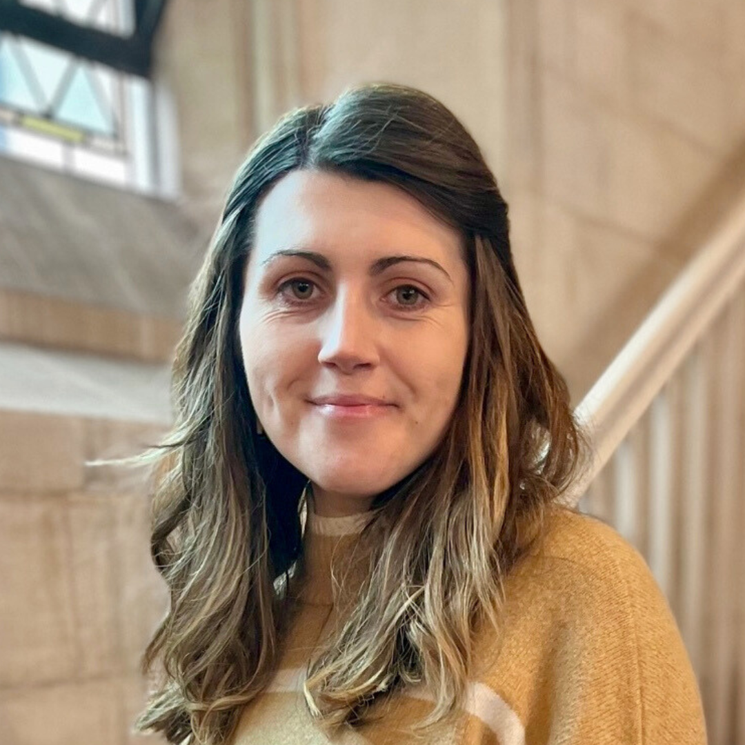 A young woman with wavy brown hair and light skin smiles at camera indoors with a wooden staircase and a window in the background.