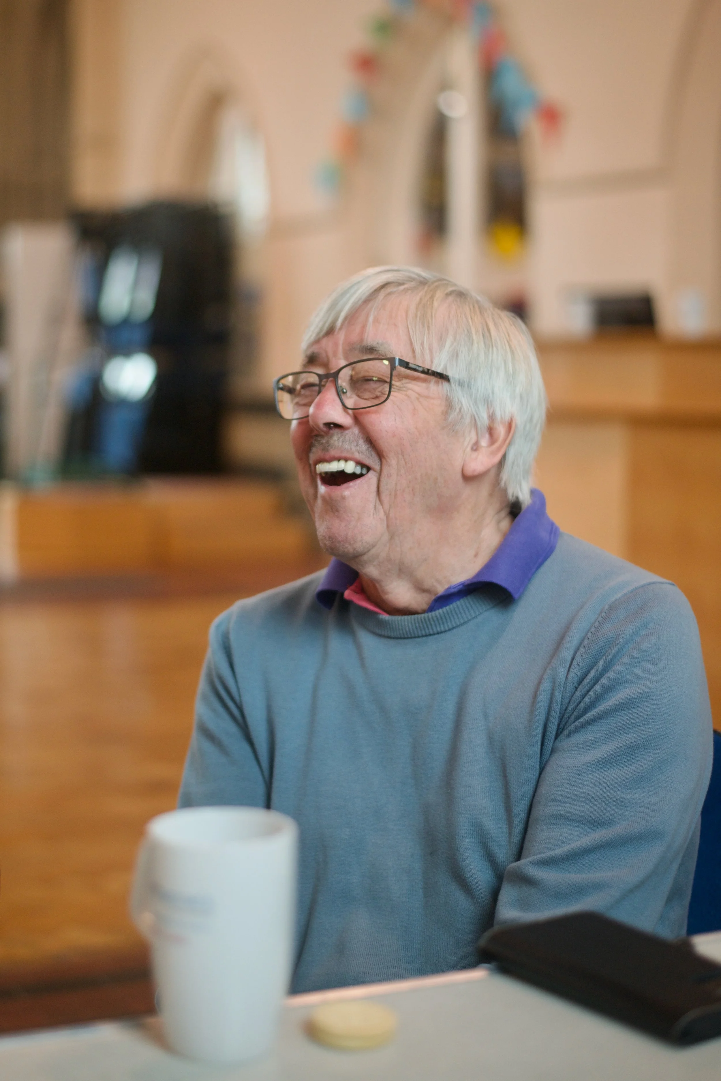 An elderly man with glasses and gray hair laughing, seated at a table with a white mug and a coin, in a cozy indoor setting.