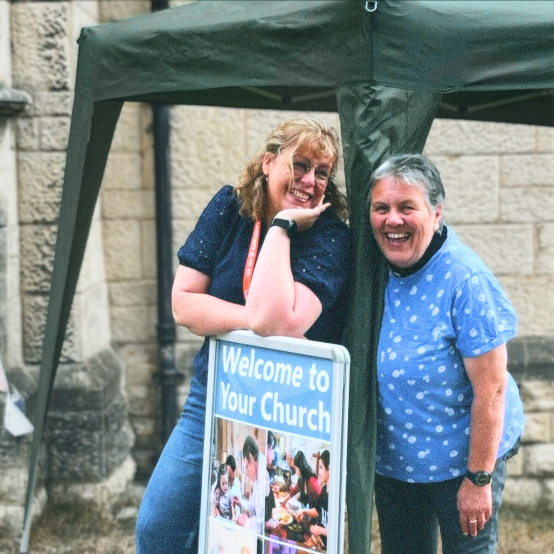 Two women smiling and posing under a green canopy next to a sign that reads 'Welcome to Your Church' with pictures of people at a community event.