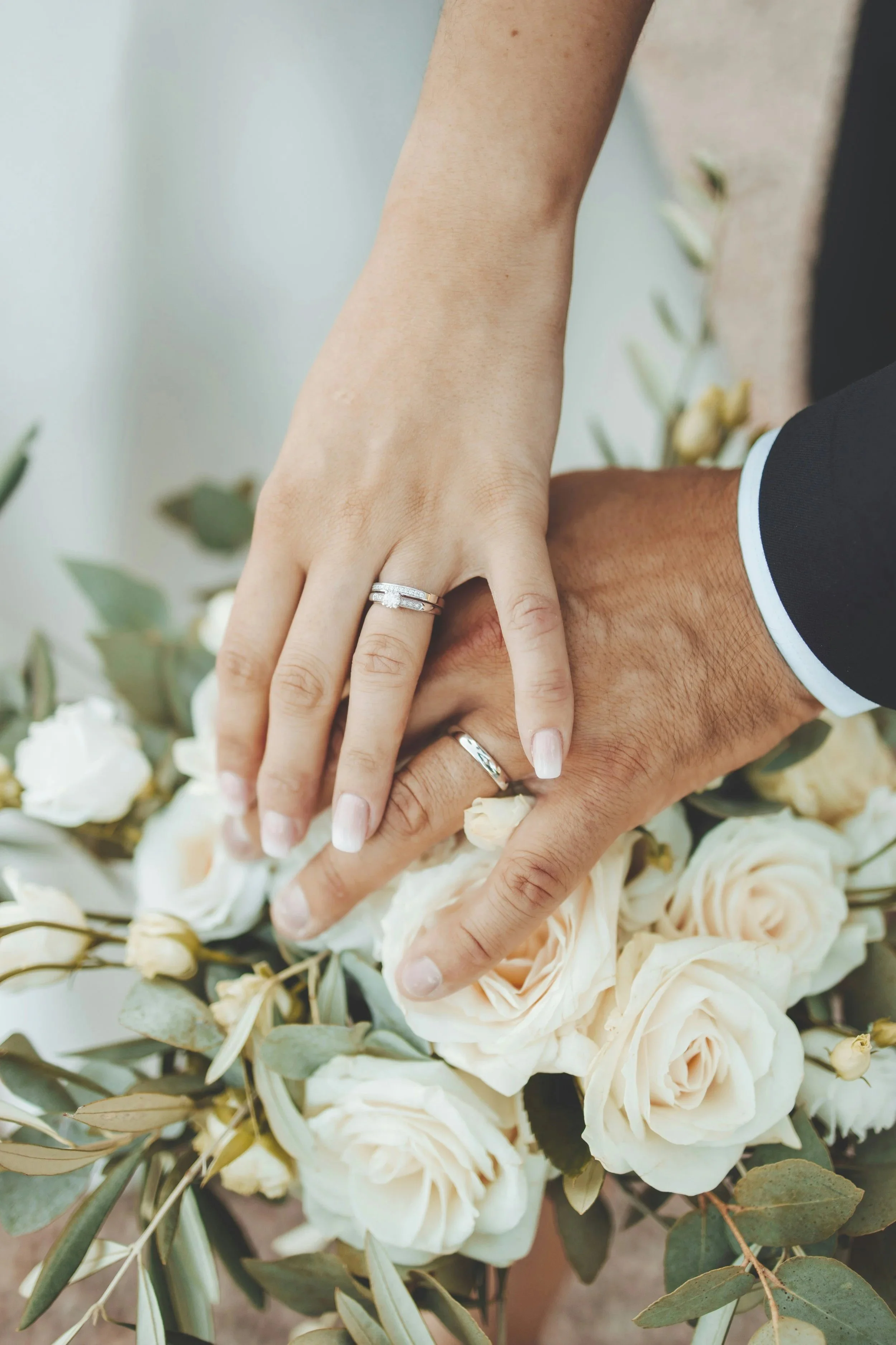 Close-up of a couple's hands with wedding rings, touching a bouquet of white roses and greenery.