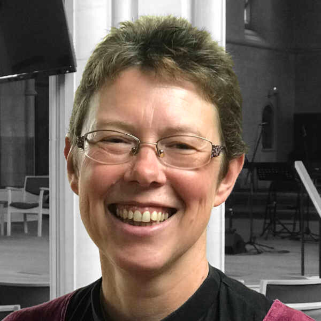 A smiling woman with short brown hair and glasses, wearing a black shirt, in an indoor setting with chairs and musical equipment in the background.
