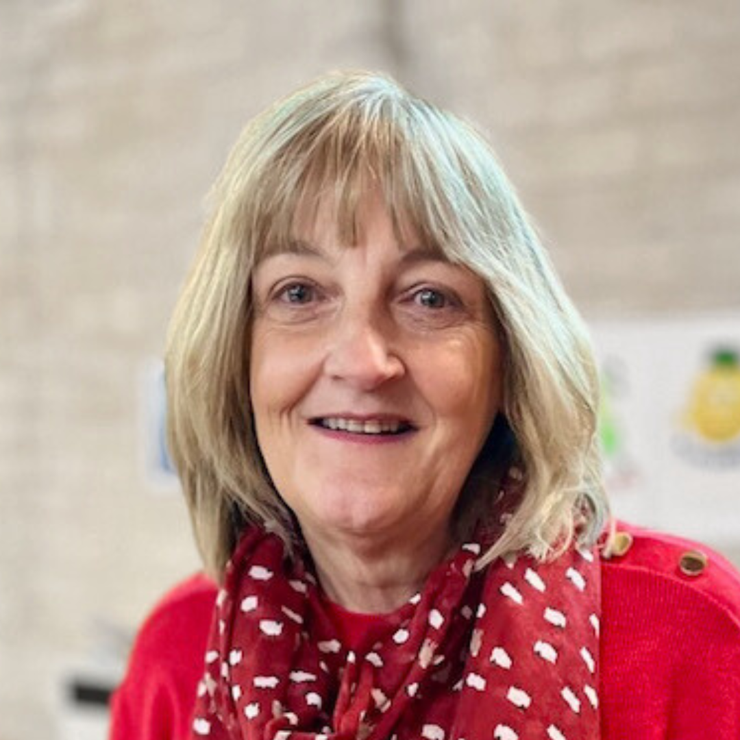 A smiling woman with blonde hair, wearing a red sweater and a red scarf with white patterns, standing indoors.