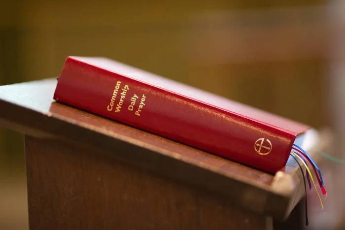 A red hardcover book titled 'Common Worship Daily Prayer' resting on a wooden surface in a church or chapel setting.