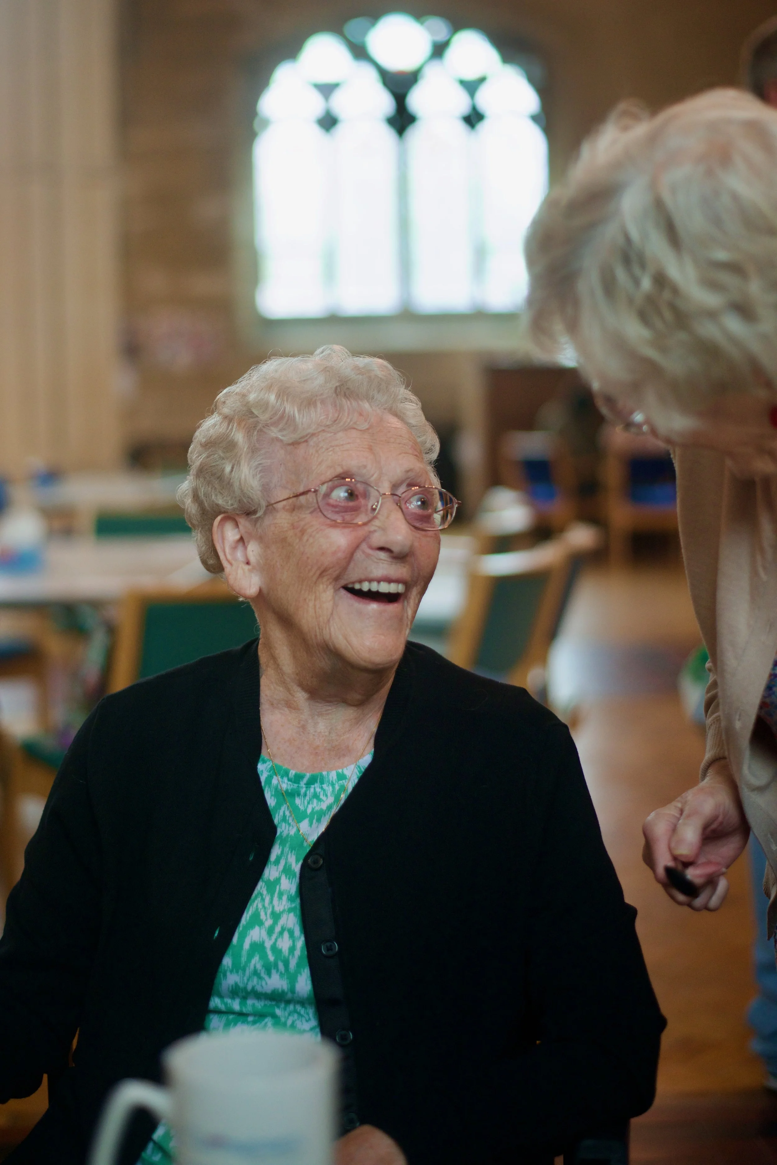 An elderly woman with short gray hair, glasses, and wearing a black cardigan and a teal patterned shirt, smiling and engaging in conversation with another woman in a bright, spacious room with large arched window and wooden furniture.