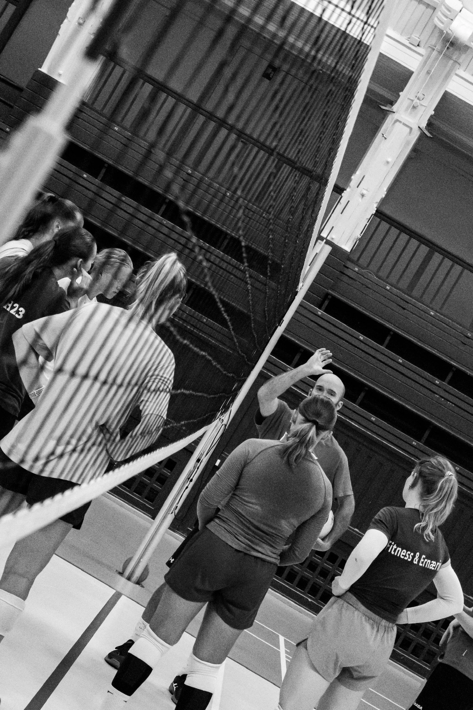 A group of women, possibly athletes, listening to a coach or trainer during a volleyball practice or game in an indoor gymnasium. The women are standing near a volleyball net, with some wearing sports attire.