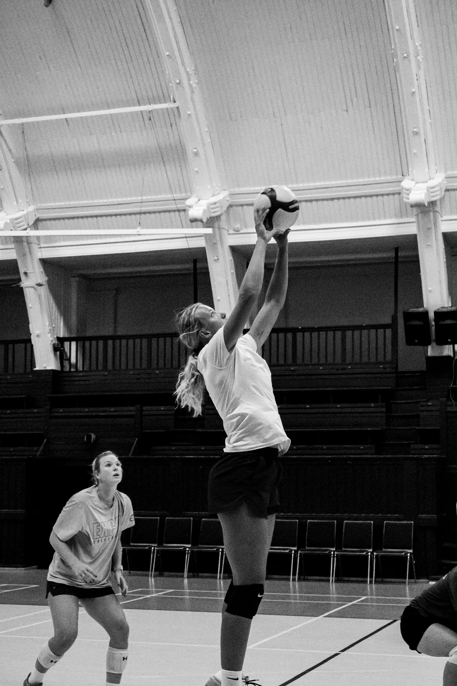 A volleyball player jumping to hit the ball during a game in an indoor gym, with another player watching nearby.