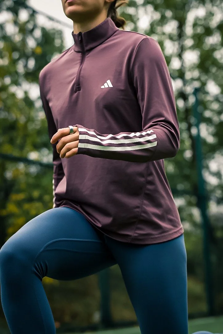 A woman in purple athletic jacket and blue leggings outdoors, stretching or warming up in a park setting.