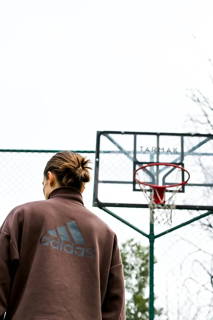 Person wearing a brown Adidas hoodie standing near a basketball hoop outdoors on a cloudy day.