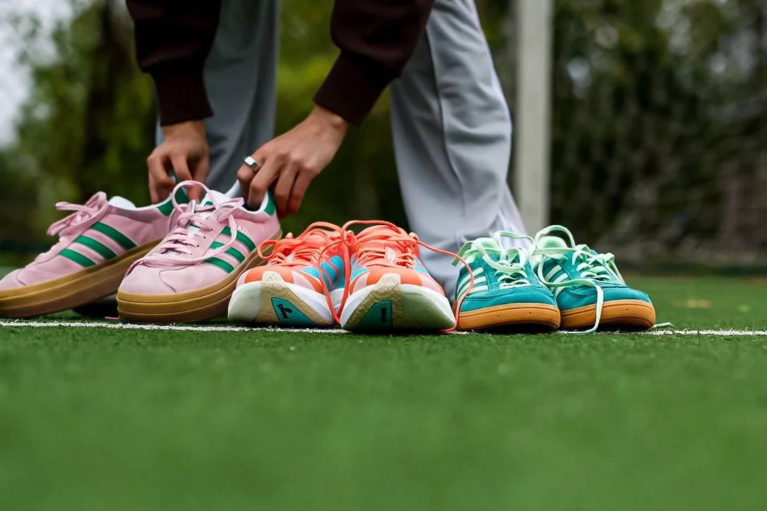 Person's hands tying pink and green sneakers on a green sports field with other colorful sneakers on the ground.
