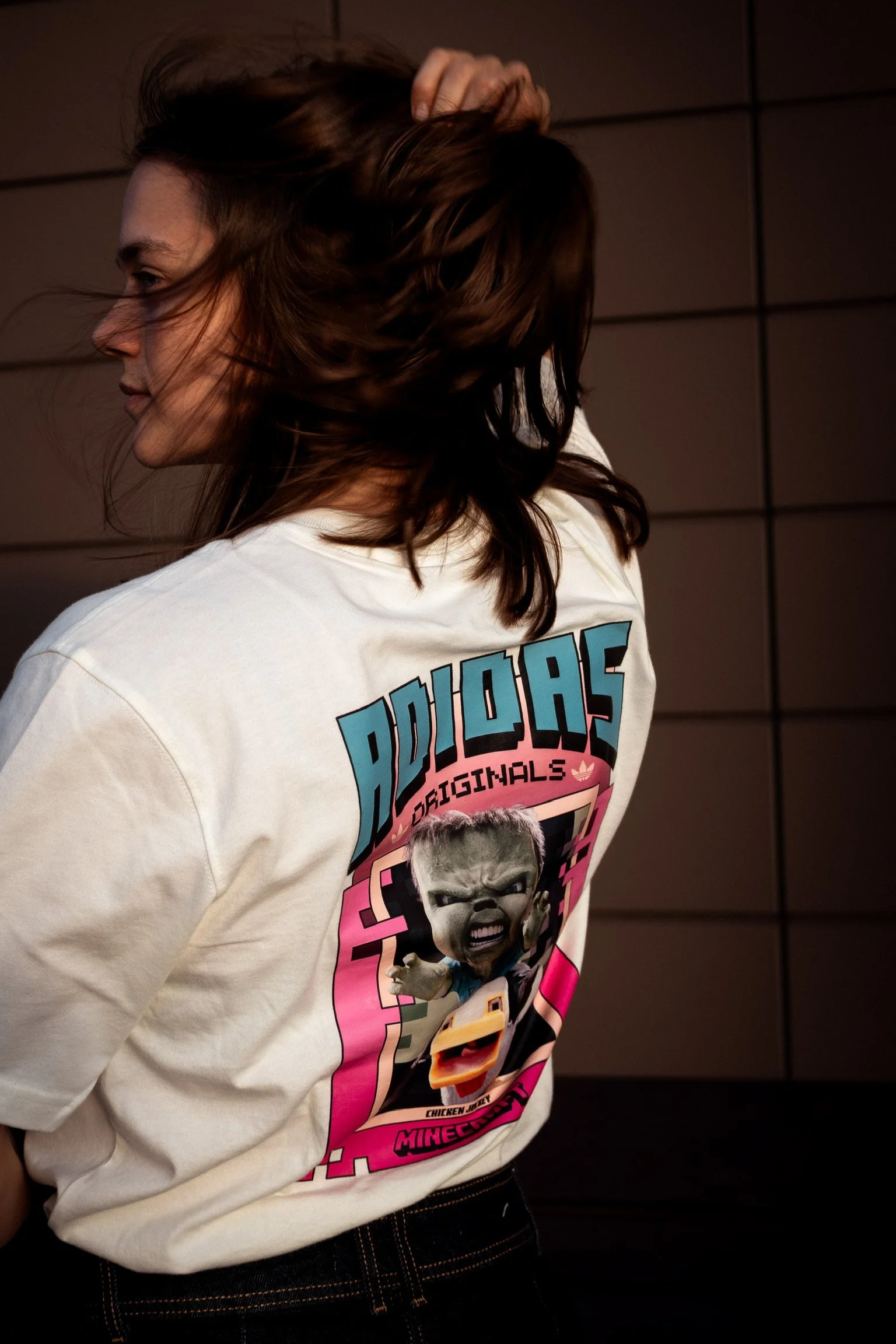 A woman with brown hair is wearing a white ADIDAS Originals T-shirt with a graphic of a crying baby with a reddish face and a beige head on the back. The woman is standing against a dark wall, with her left arm raised and hand in her hair, partially 