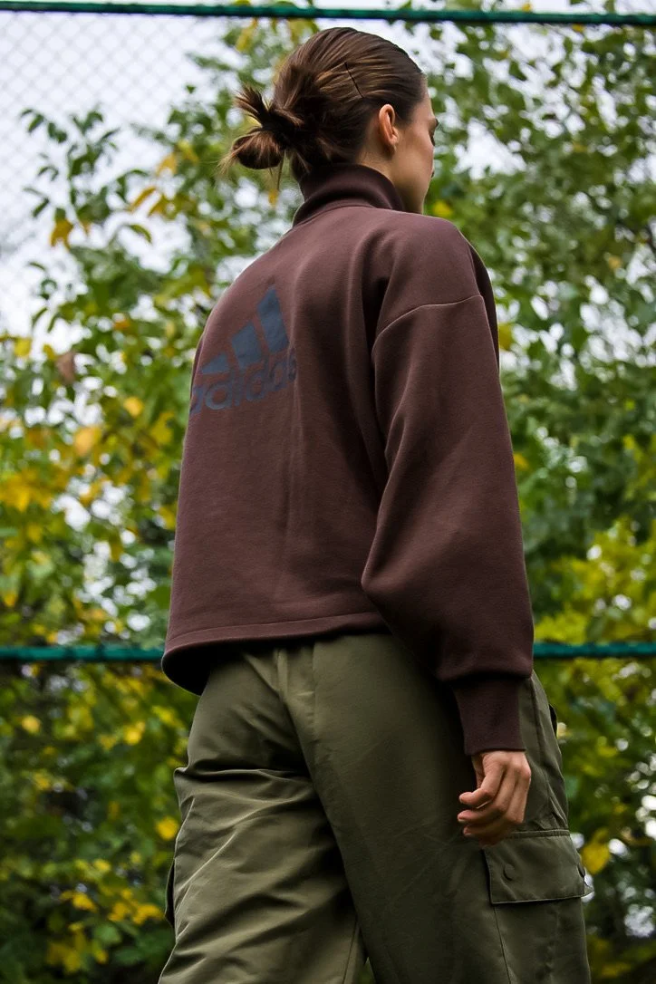 A woman seen from behind wearing a brown Adidas sweatshirt and olive green cargo pants outdoors with trees in the background.