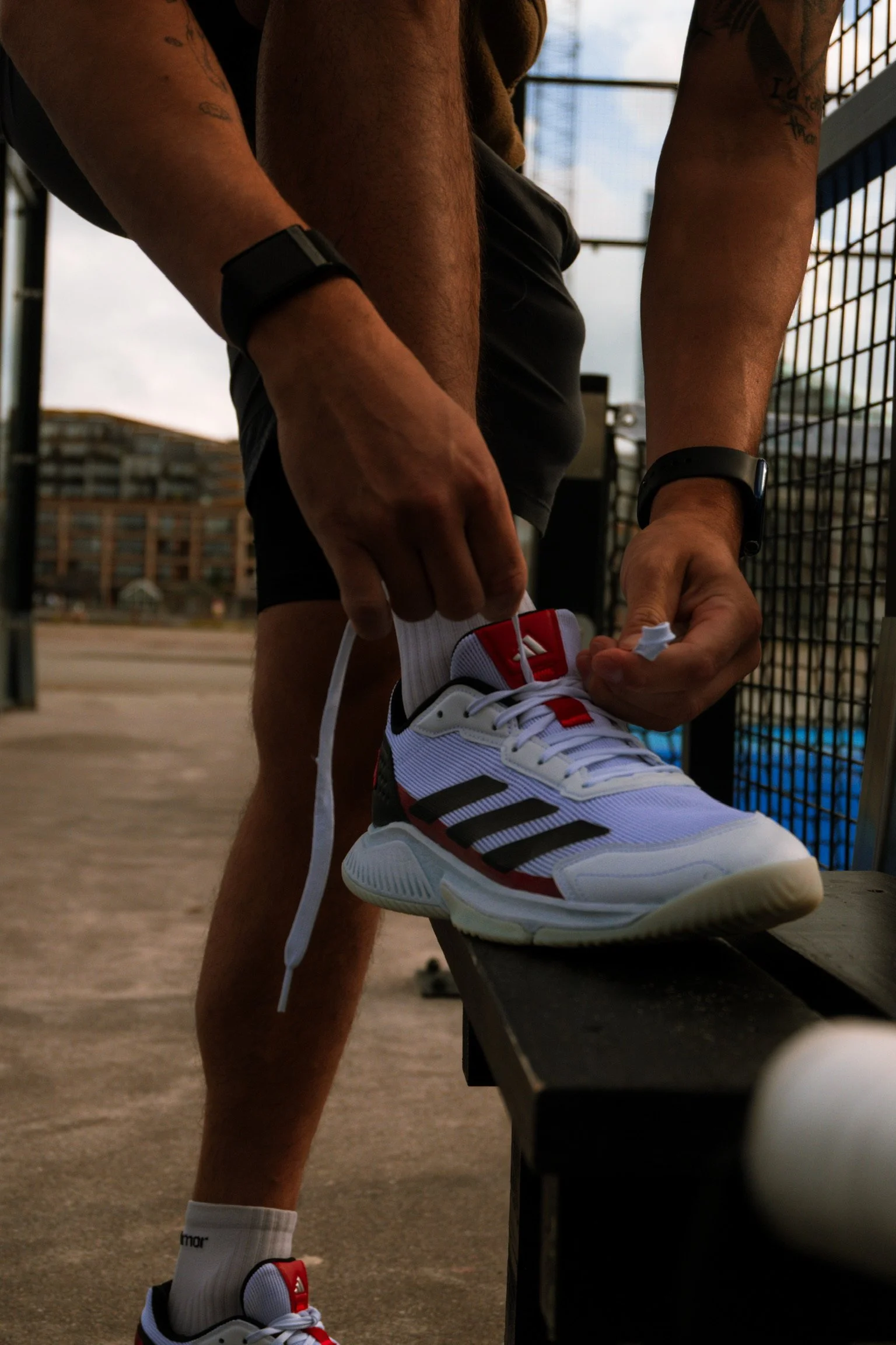 A person is tying the laces of a white athletic shoe with red and black details, standing on a bench outdoors.