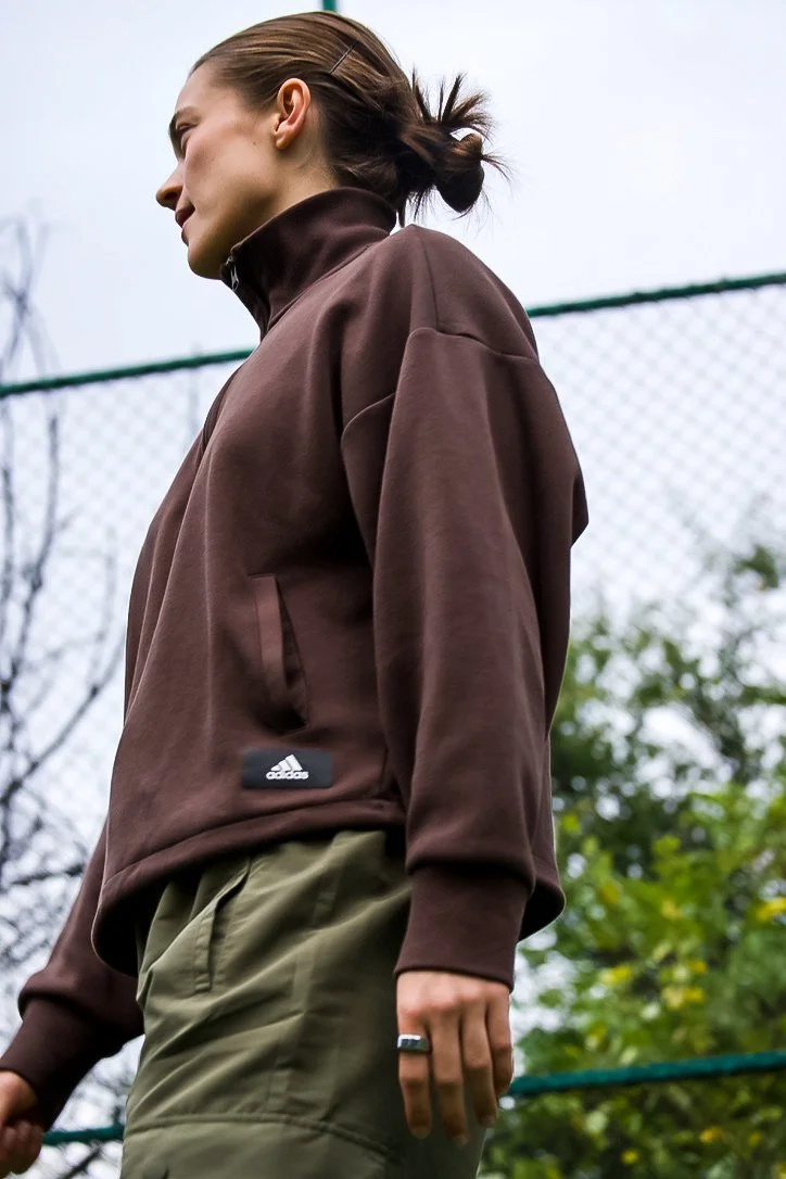 A woman standing outdoors near a chain-link fence, wearing a brown jacket and khaki pants, with her hair tied back in a bun.