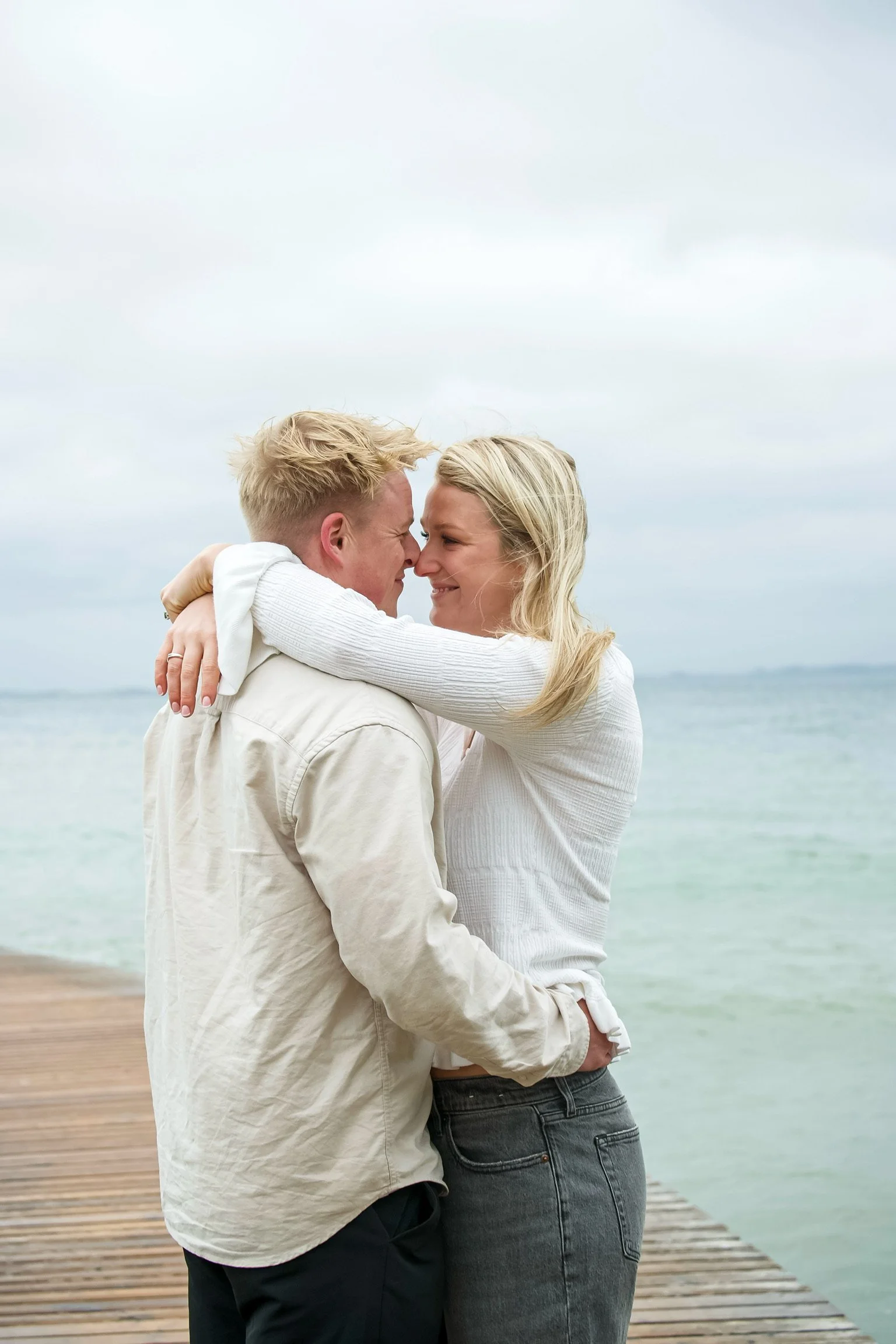 A couple hugging on a wooden pier by the water, smiling at each other with noses touching.