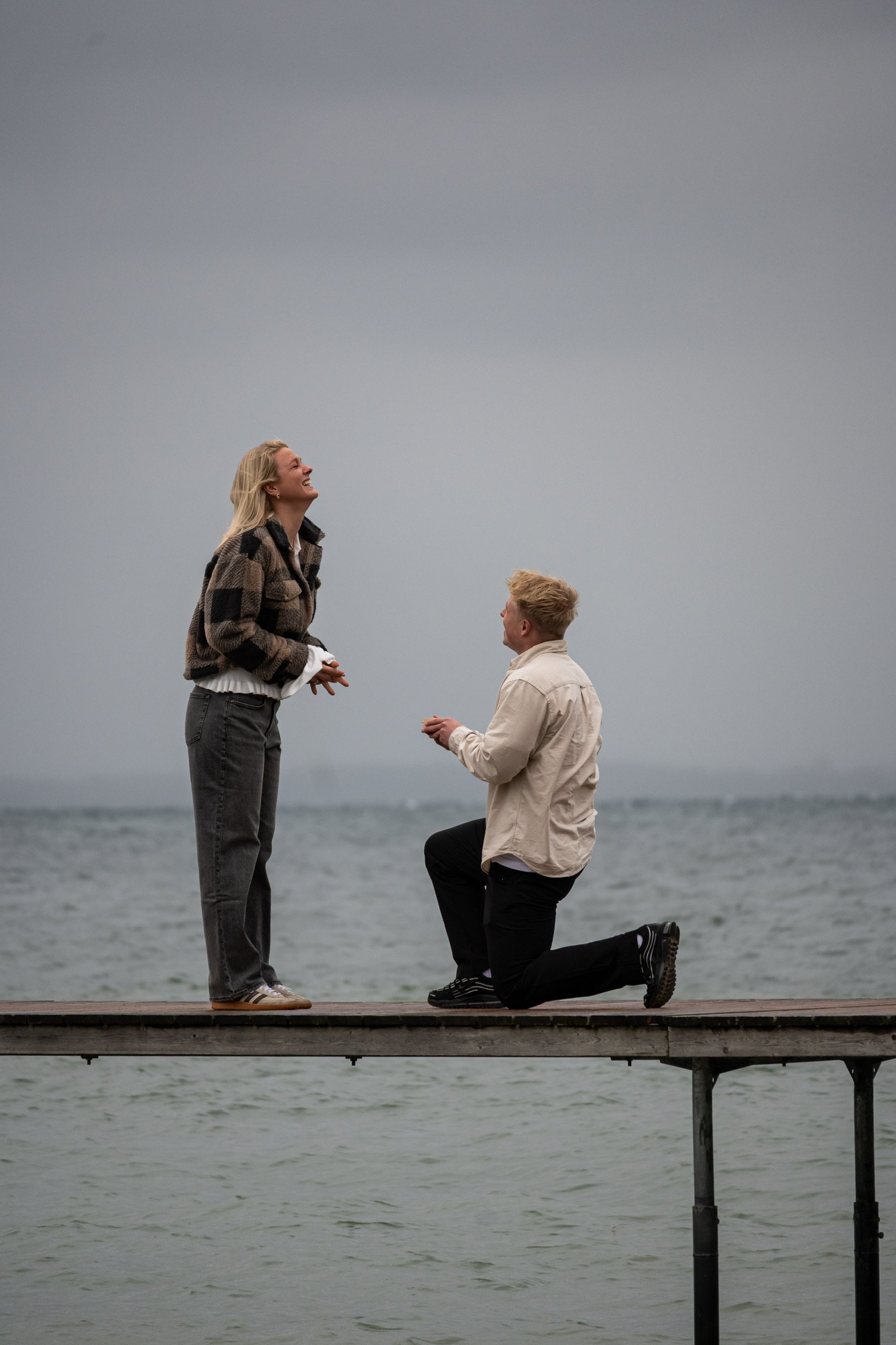A man kneeling on one knee on a dock by the water, proposing to a woman standing in front of him, both smiling. Overcast sky in the background.