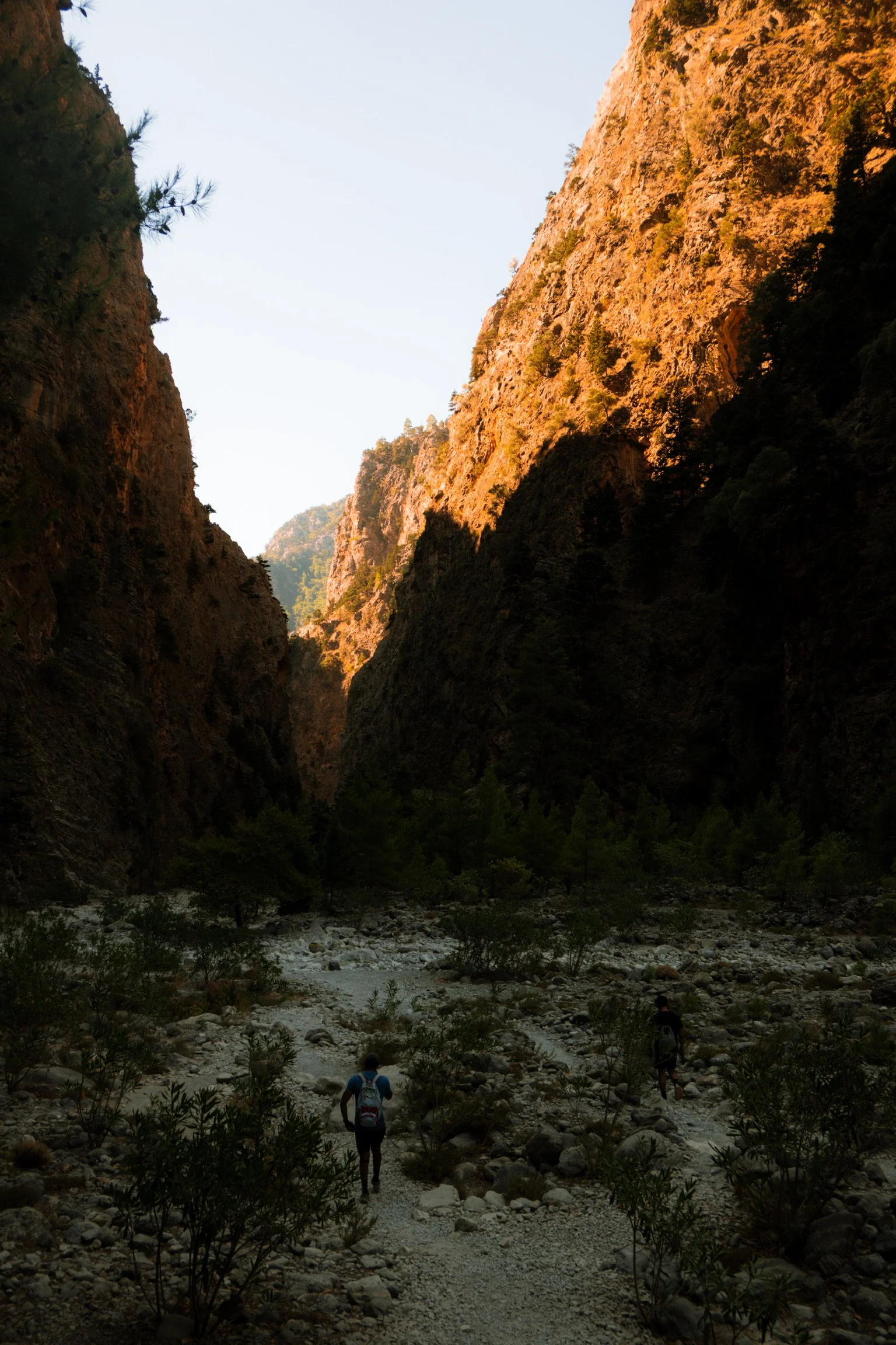 Two hikers walking through a rocky canyon with steep, sunlit cliffs on either side.