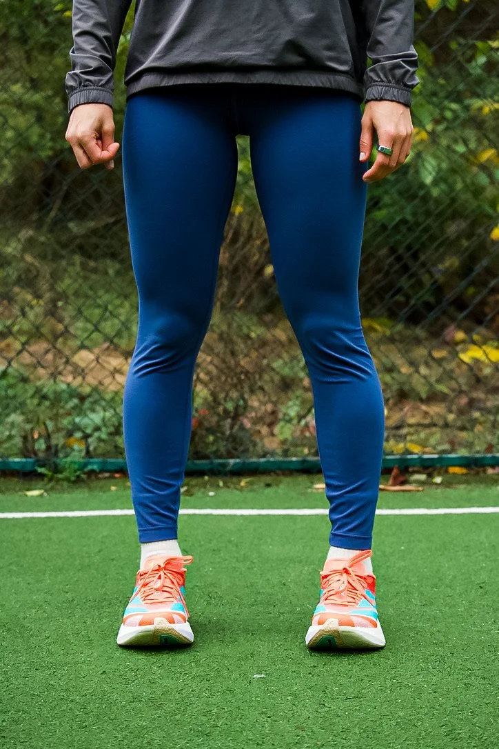 A person standing on a sports field wearing a gray jacket, blue athletic leggings, and colorful running shoes.
