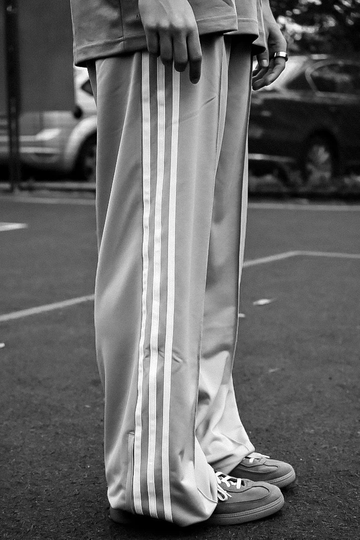 Close-up of a person's lower body wearing loose-fitting pants with three vertical stripes down the side, sneakers, and a ring on their finger, standing on a parking lot.