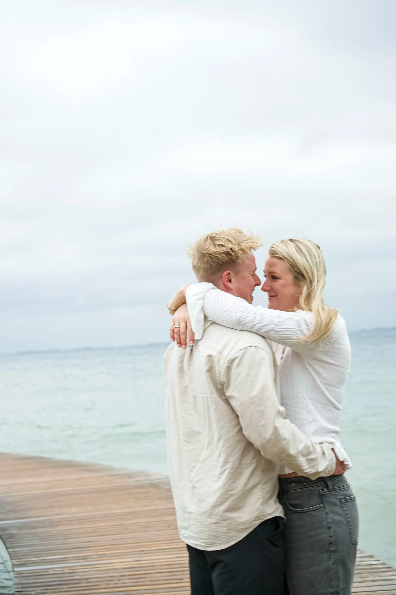 A couple hugging on a dock by the ocean with an overcast sky.