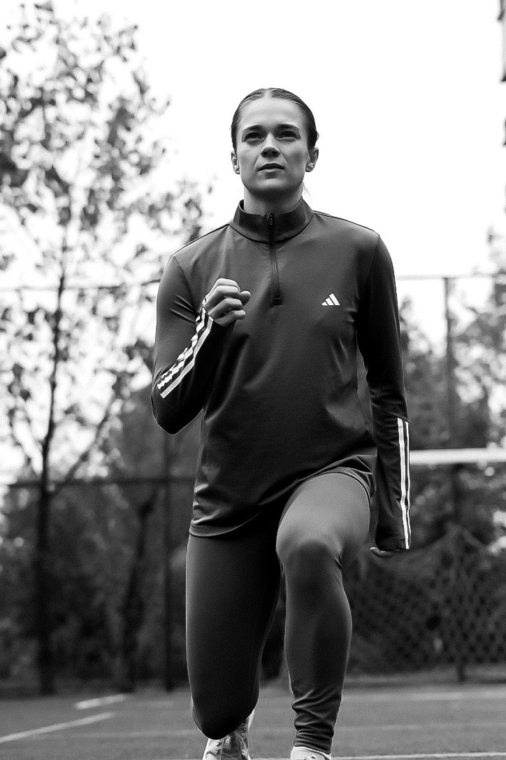 A woman running outdoors on a tennis court, wearing a sporty Adidas jacket and leggings, with trees and a fence in the background.