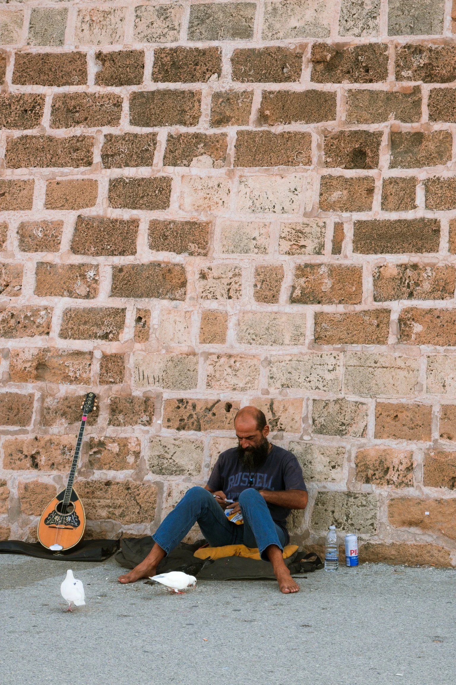 A man with a beard sitting barefoot on the street against a large brick wall. There is an acoustic guitar leaning against the wall, and he is surrounded by a water bottle, a can, and a cardboard box. Two white birds are near him, one on the ground an
