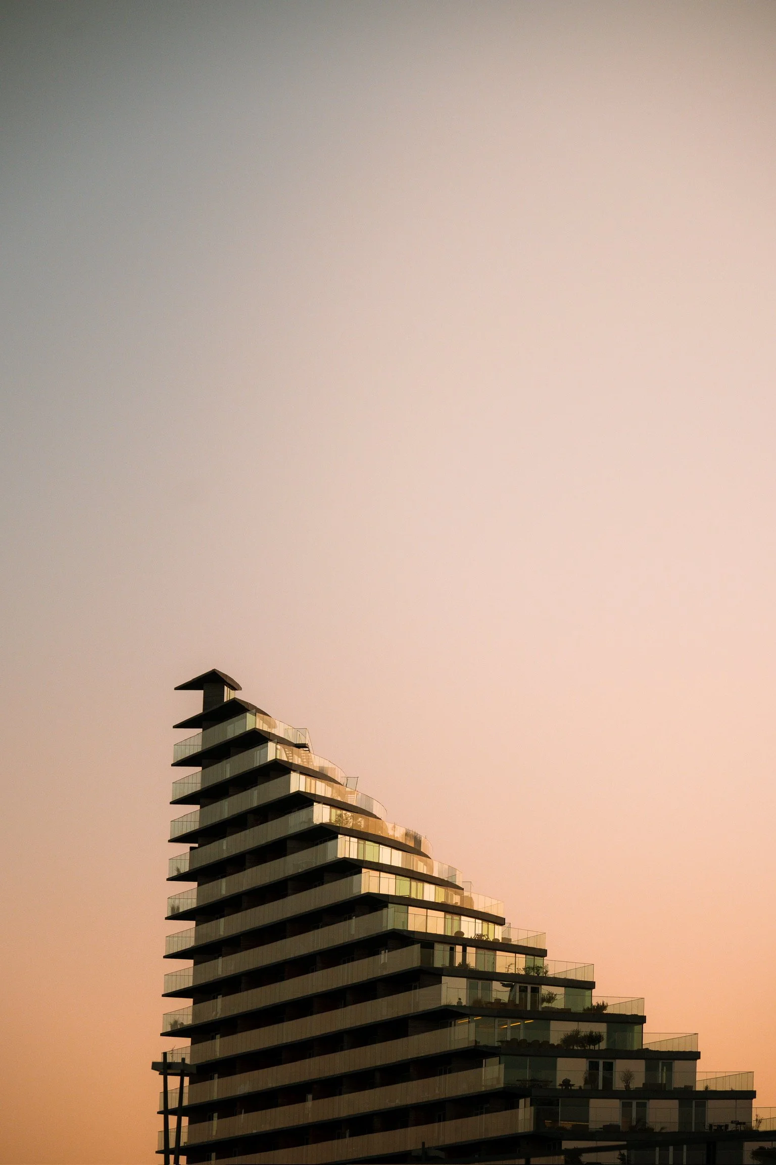 A modern multi-story building with glass balconies, tapering in a stepped design, against a gradient sky at sunset.