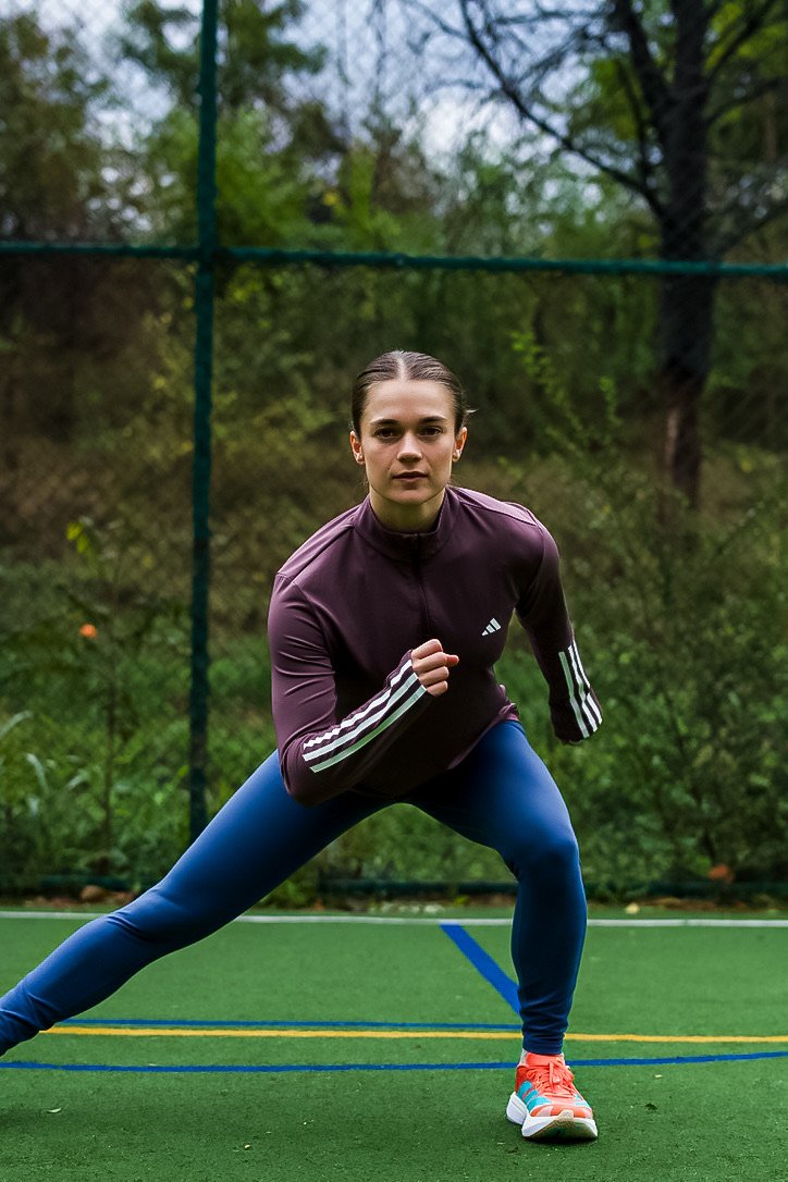 Young woman in athletic clothing stretching on outdoor sports court with trees in background.