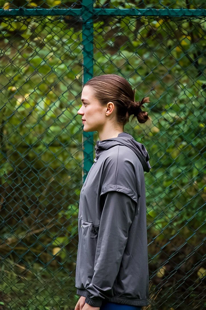 Side view of a woman standing outdoors near a chain-link fence with green foliage in the background, wearing a gray jacket and her hair tied back.