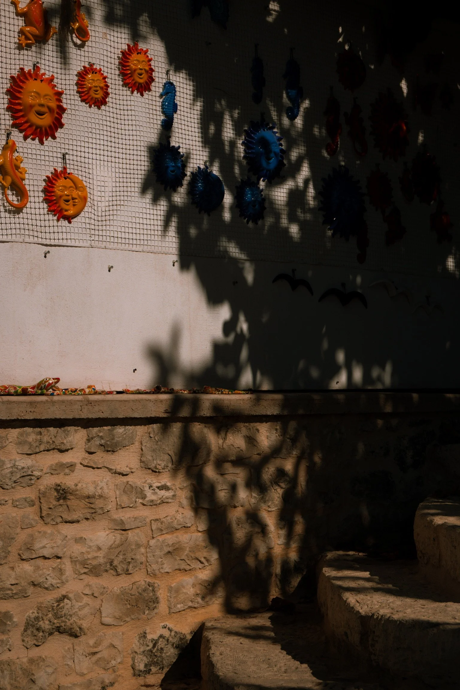 Colorful ceramic sun and seahorse decorations hanging on a wire mesh display with shadows cast on a white wall and stone steps below.