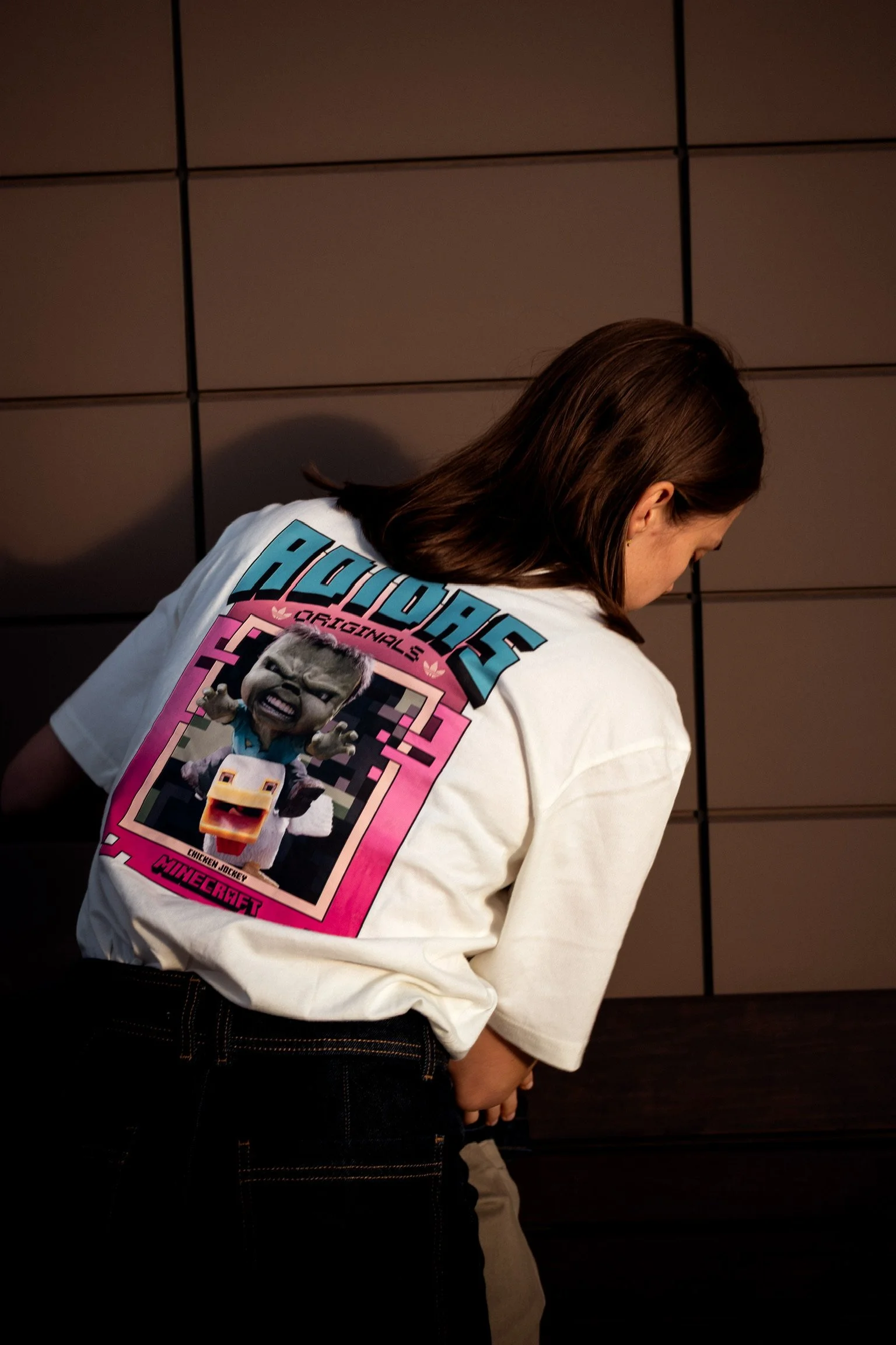 Person with brown hair wearing a white t-shirt with a colorful graphic on the back featuring characters from Minecraft and other pop culture references, standing against a dark wall.