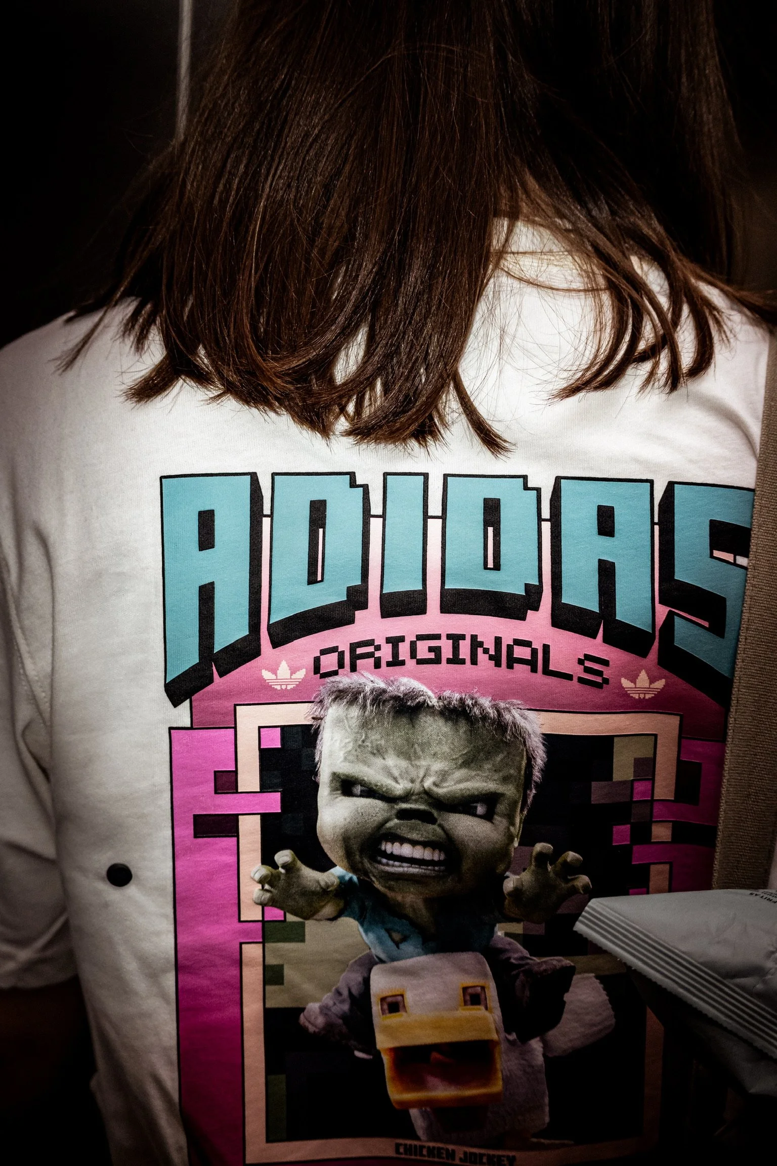 Close-up of a person wearing a white T-shirt with a graphic design featuring a cartoon character with an angry face and a duck bill. The design includes the word 'ADIDAS' in pixelated style and the phrase 'ORIGINALS' with the Adidas logo.