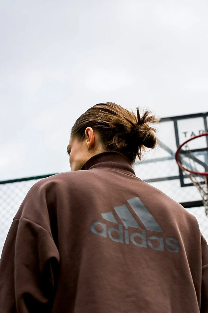 Person with brown hair tied in a bun wearing a brown Adidas jacket on a basketball court.