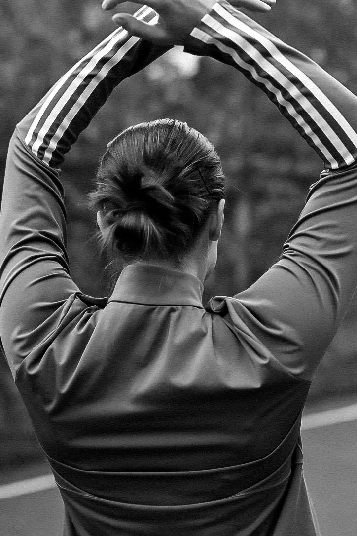 A woman with her hair in a bun, wearing a sports jacket with striped sleeves, stretching outdoors with her arms raised above her head.