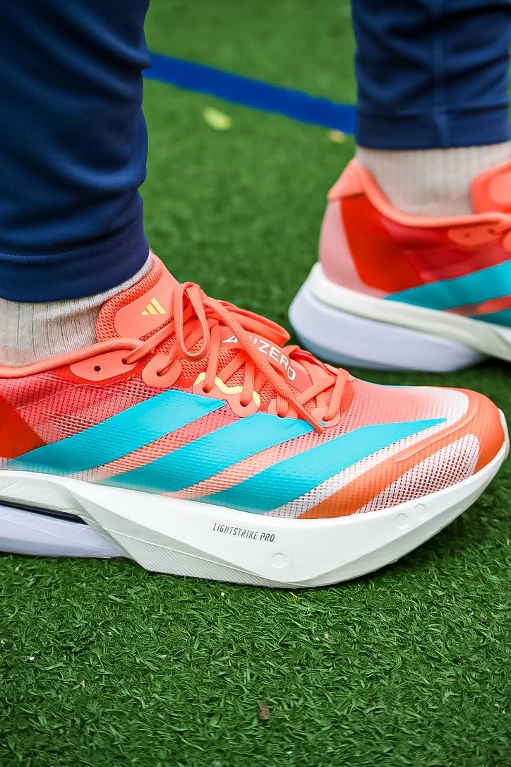 Close-up of coral and turquoise Nike running shoes on green turf, worn with navy pants and beige socks.