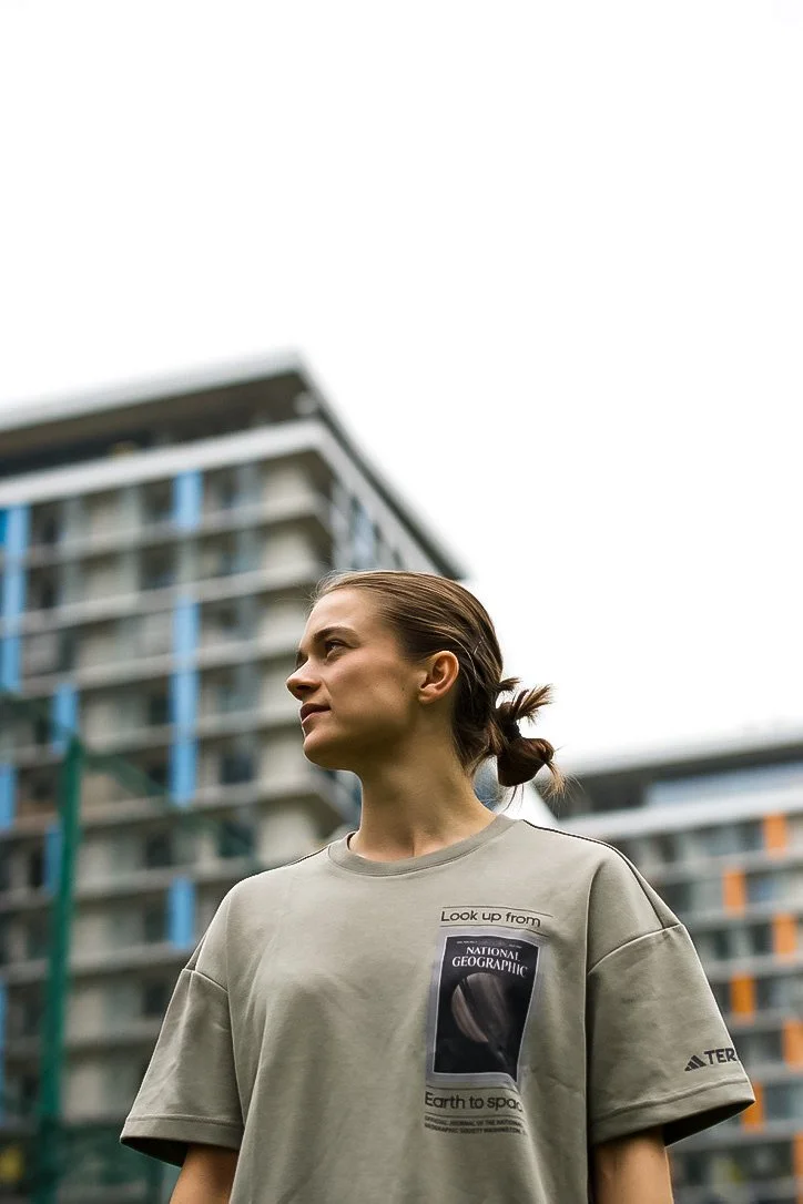 Young woman with brown hair tied in a small ponytail standing outdoors in front of a modern multi-story building, wearing a gray National Geographic t-shirt, looking to the side.
