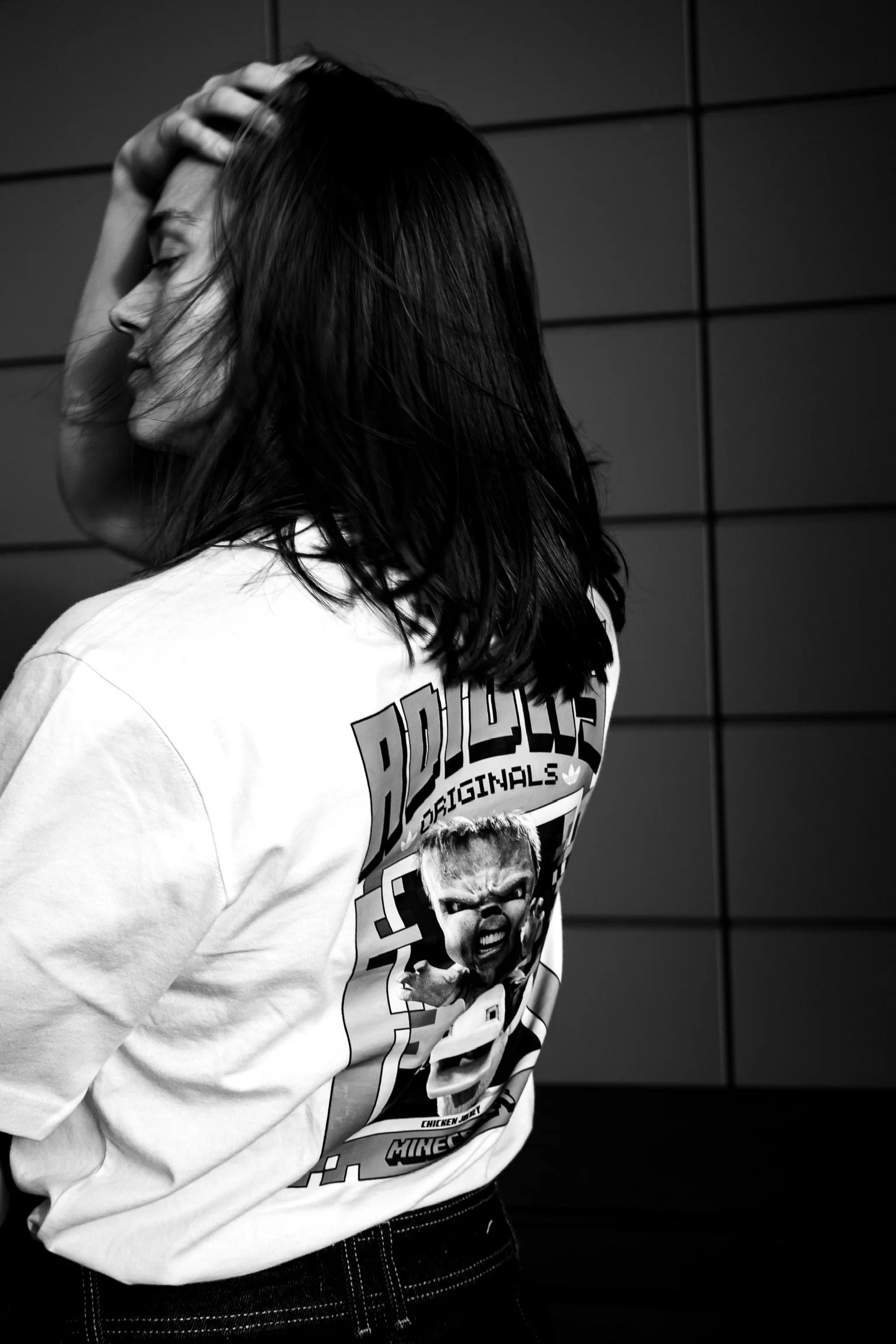 A woman with shoulder-length dark hair, wearing a graphic t-shirt with a cartoon character, smiling and leaning against a wall with horizontal panels, in black and white.