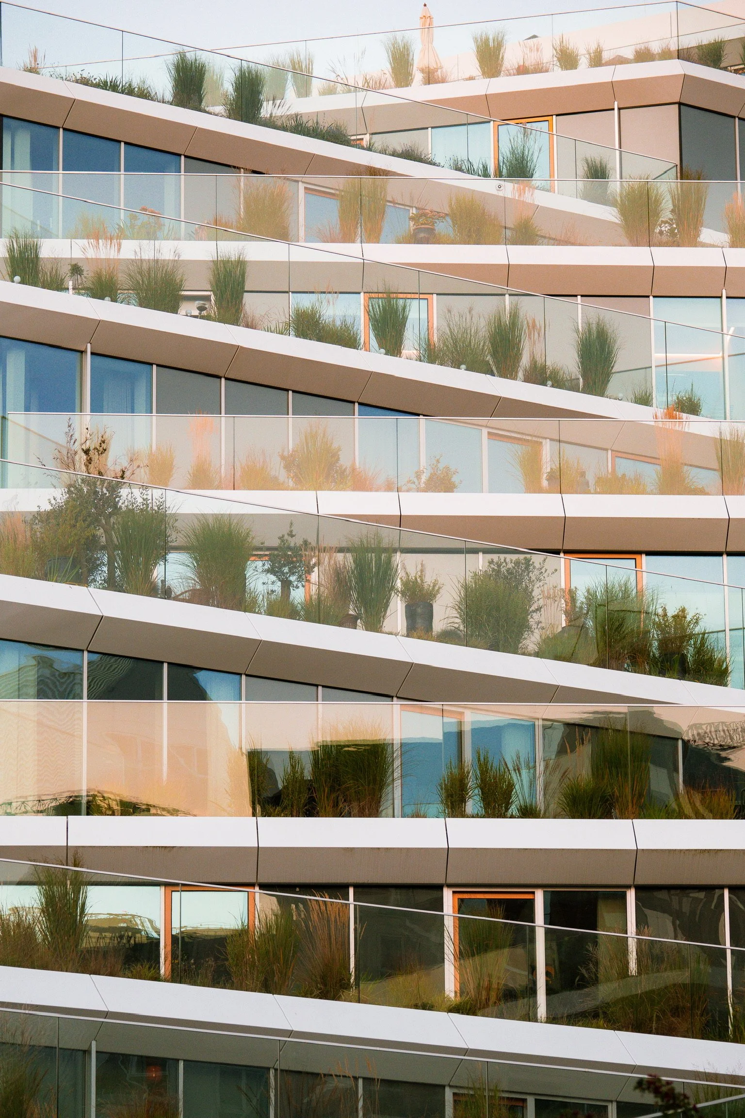 Modern multi-story building with large glass windows and balconies decorated with potted plants and greenery, reflecting the sky and surroundings.
